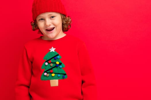 cheerful funny boy on a red background in a warm hat and sweater with a Christmas tree.