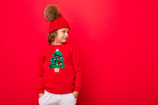 cool teenager in a red Christmas sweater fooling around against the background of a red wall, a warm hat and a sweater with a Christmas tree.