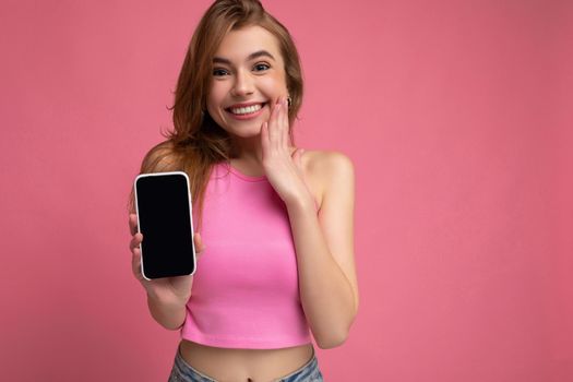 Photo of beautiful smiling young woman good looking wearing casual stylish outfit standing isolated on background with copy space holding smartphone showing phone in hand with empty screen display for mockup looking at camera.