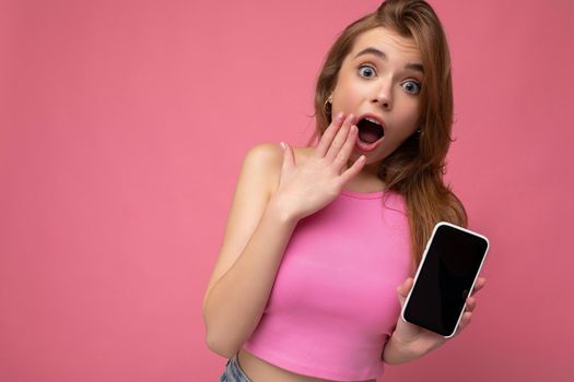 Photo of amazed beautiful joyful young blonde woman wearing pink top poising isolated on pink background with empty space holding in hand and showing mobile phone with empty display for mockup looking at camera.