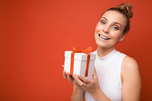 Beautiful happy young blonde woman isolated over colourful background wall wearing stylish casual clothes holding gift box and looking at camera. Empty space