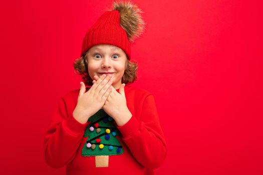 cool teenager in a red Christmas sweater fooling around against the background of a red wall, a warm hat and a sweater with a Christmas tree.