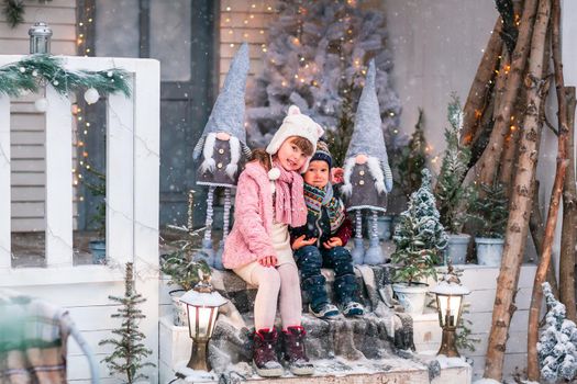 Happy little kids sitting on the porch of the Christmas decorated house, snowing outdoor. Happy New Year and Merry Christmas. Magic winter