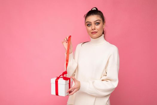 Attractive positive surprised young brunette woman isolated over pink background wall wearing white sweater holding gift box and unboxing present looking at camera.