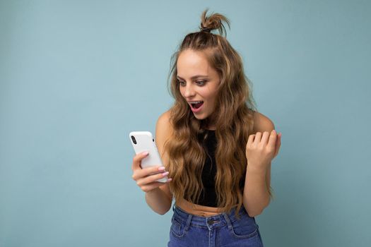 Charming positive young blonde woman wearing black top poising isolated on blue background with empty space holding in hand and using mobile phone communicating online looking at gadjet.