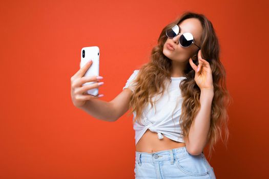 Attractive charming young smiling happy woman holding and using mobile phone taking selfie wearing stylish clothes isolated over wall background.