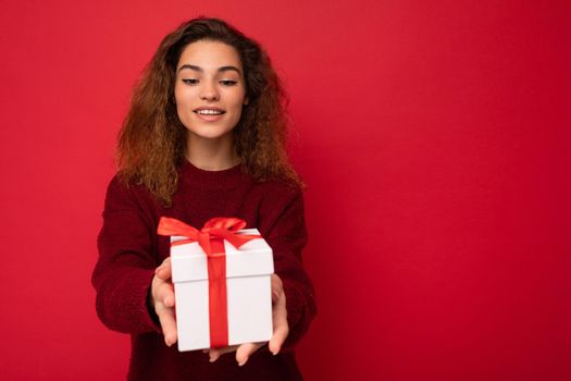 Attractive positive smiling young brunette curly woman isolated over red background wall wearing red sweater holding gift box looking at present white box with red ribbon. Empty space