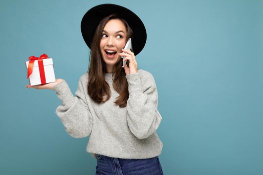 Beautiful happy young brunette woman isolated over colourful background wall wearing stylish casual clothes holding gift box and looking to the side.