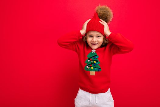 cheerful funny boy on a red background in a warm hat and sweater with a Christmas tree.