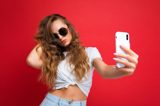 Closeup of sexy amazing beautiful young woman holding mobile phone taking selfie photo using smartphone camera wearing round sunglasses everyday stylish outfit isolated over colorful wall background looking at device screen.