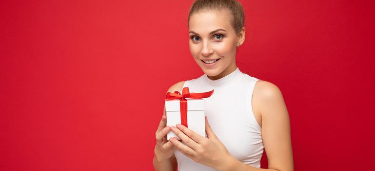 Shot of beautiful positive smiling young blonde woman isolated over red background wall wearing white top holding gift box and looking at camera. Copy space, mockup