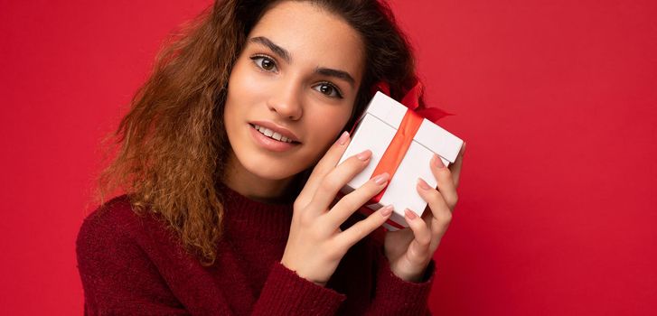 Closeup photo of attractive happy joyful young brunette curly female person standing isolated on red background wall wearing red sweater holding gift box with red ribbon looking at camera.