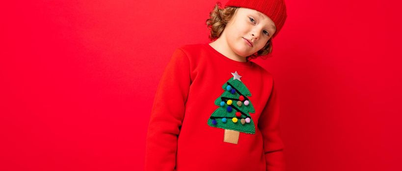 cool teenager in a red Christmas sweater fooling around against the background of a red wall, a warm hat and a sweater with a Christmas tree.