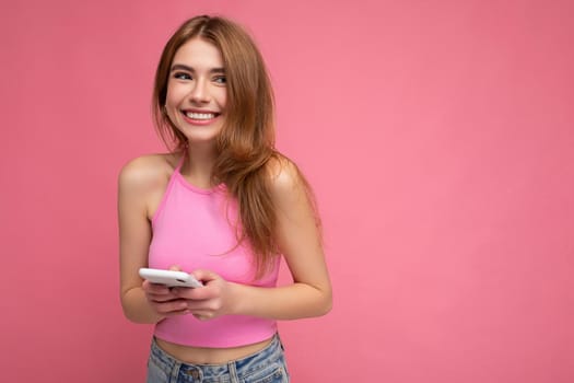 Photo of smiling good looking young blonde woman wearing pink top poising isolated on pink background with empty space holding in hand and using mobile phone messaging sms looking to the side. copy space
