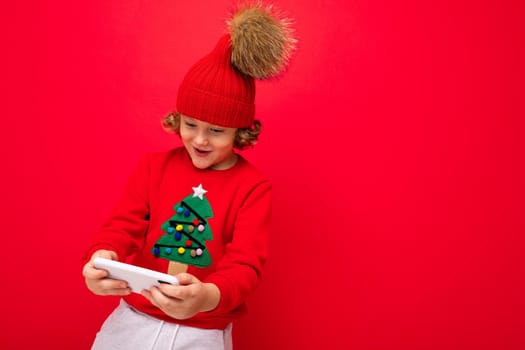 a young man with a smartphone in his hands against the background of a red wall, in a knitted hat and a sweater with a Christmas tree