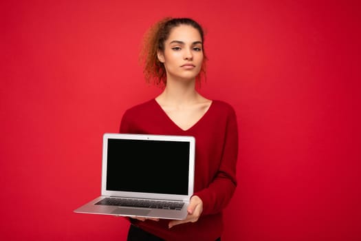 Close-up portrait of beautiful dark blond curly young girl holding laptop computer looking at camera wearing red sweater isolated over red wall background.