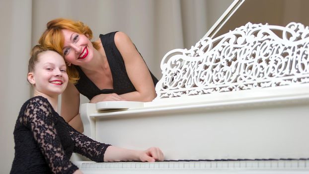 A beautiful young mother with a little daughter near a large white grand piano. The concept of family happiness, music education.