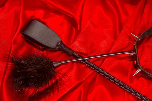 Bondage, kinky adult sex games, kink and BDSM lifestyle concept with a whip, feather stick, collar on red silk with copy space