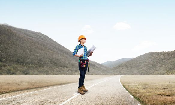 Young woman in safety helmet standing on asphalt road with blueprints. Female architect in workwear inspection road construction on background of hills. Transportation engineering and construction.