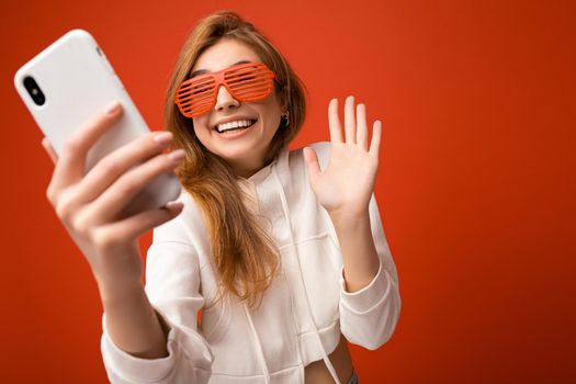 Attractive charming young smiling happy woman holding and using mobile phone taking selfie wearing stylish clothes isolated over wall background.