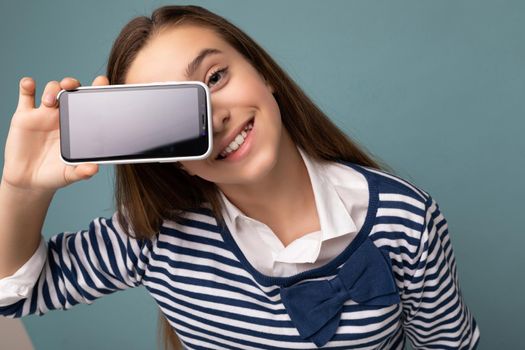Photo of beautiful smiling girl good looking wearing casual stylish outfit standing isolated on background with copy space holding smartphone showing phone in hand with empty screen display for mockup looking at camera.