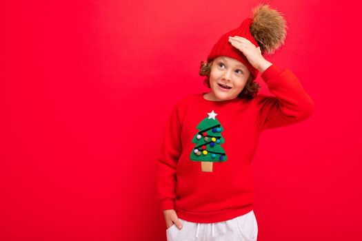 cheerful funny boy on a red background in a warm hat and sweater with a Christmas tree.