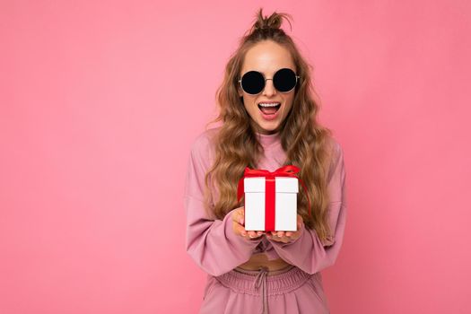 Beautiful happy young brunette woman isolated over colourful background wall wearing stylish casual clothes holding gift box and looking at camera. Copy space