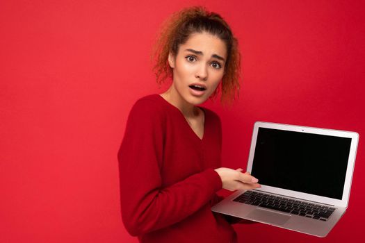 Close-up portrait of beautiful amazed surprised astonished dark blond woman holding laptop computer looking at camera showing at netbook keyboard wearing red sweater isolated over red wall background.