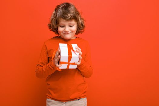 Positive blonde curly boy isolated over red background wall wearing red sweater holding gift box and looking at surprise box. Empty space