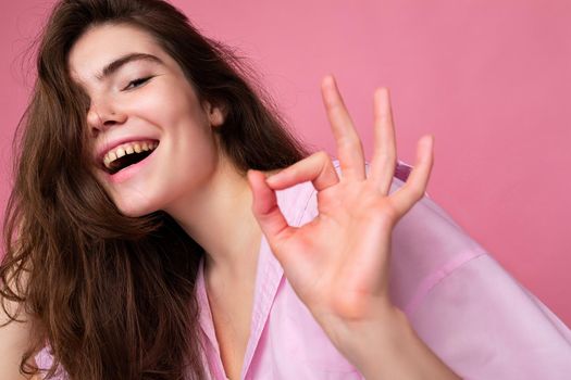 Closeup of young positive happy smiling charming sexy curly brunette woman with sincere emotions wearing trendy pink shirt isolated on pink background with empty space and showing ok gesture. It's good.