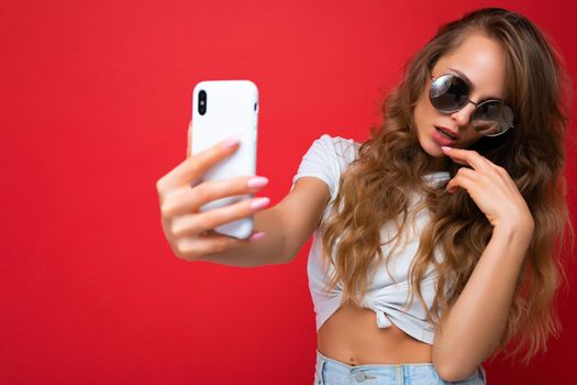 Closeup photo of amazing beautiful young blonde woman holding mobile phone taking selfie photo using smartphone camera wearing sunglasses everyday stylish outfit isolated over colorful wall background looking at device screen.