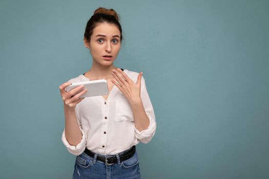 Pretty sad young blonde curly woman wearing casual white shirt and isolated over blue background wall holding and using phone looking at camera. copy space
