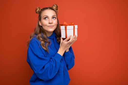 Beautiful happy young blonde woman isolated over colourful background wall wearing stylish casual clothes holding gift box and looking to the side. Copy space