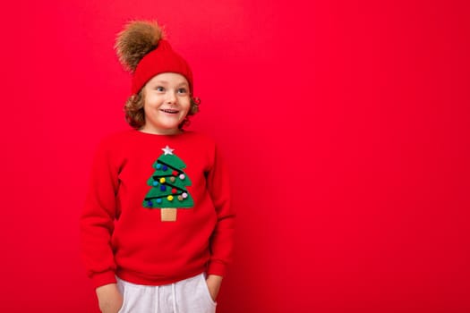 cheerful funny boy on a red background in a warm hat and sweater with a Christmas tree.