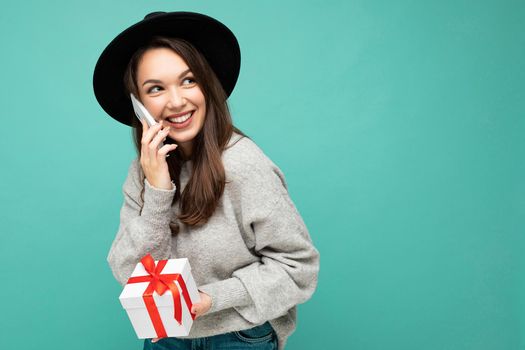 Photo of beautiful happy positive joyful young brunette woman isolated over blue background wall wearing black hat and grey sweater holding gift box talking on mobile phone and looking to the side. Copy space, mockup