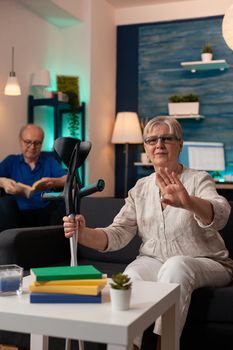 Retired woman with crutches waving on video call conference at home. Older wife using online remote communication while disabled husband sitting in wheelchair in living room background