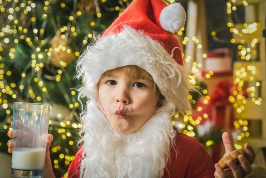 Little funny Santa child eating Christmas cookie and glass of milk on Christmas tree background