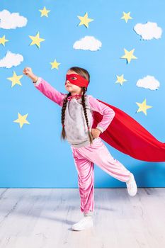 Six year blonde girl dressed like superhero having fun at home. Kid on the background of bright blue wall with white clouds and yellow stars. Girl power concept.