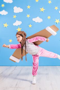 Girl pilot playing with toy jet pack at home. Success and leader concept. Kid on the background of bright blue wall with white clouds and yellow stars.