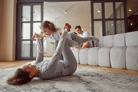 Jolly mother is doing gymnastics with girl on carpet while father and son are using notebook