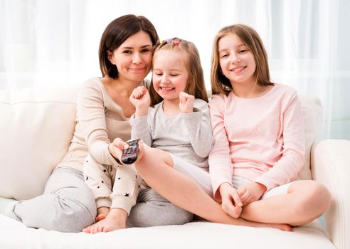 Portrait of mom hugging her cute daughters while sitting on the sofa and watching TV at home. Happy family relaxing on the coach