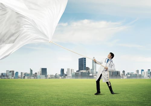 Horizontal shot of young doctor in white medical uniform pulling white fabric while standing on green meadow with cityscape view on background