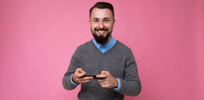 Panoramic photo shot of cool good looking brunet unshaven young man with beard wearing grey sweater and blue shirt isolated over pink background wall with empty space holding in hand and using mobile phone messaging sms online looking at camera.