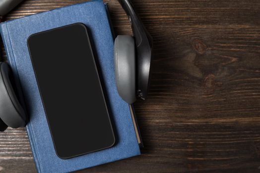 Mobile phone lying on the book with headphones. Audiobooks concept. Dark wooden background copy space