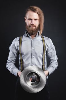 Studio portrait of a bearded stylish long-haired man in a shirt with suspenders. Holding car parts new Steel brake disc