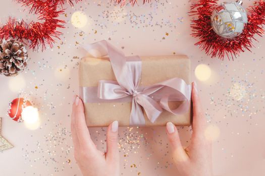 Female hands with natural manicure holding present in kraft paper with pink ribbon on soft beige background. Xmas composition. Flat lay. Happy holidays, New Year celebration and giving love concept.