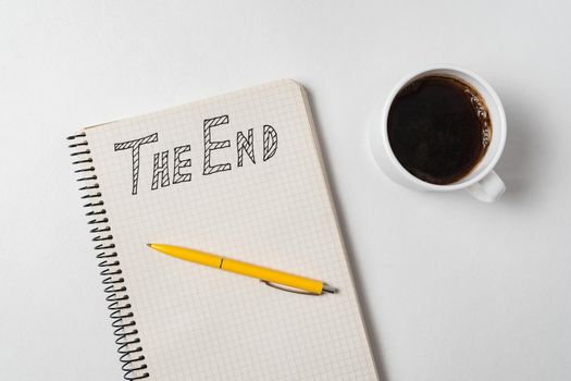 The end text on copybook. Notepad, pen and cup of coffee on white background. Top view.