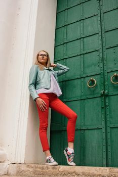 Fashionable young model girl in vintage fashion jacket with jeans and Glasses posing near green old wooden doors on the street