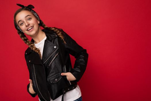 Attractive cool positive smiling brunetfemale teenager with pigtails wearing stylish black leather jacket and white t-shirt isolated over red background wall looking at camera. Copy space