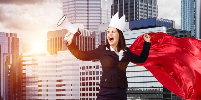 Young confident businesswoman wearing red cape against modern city background screaming in megaphone